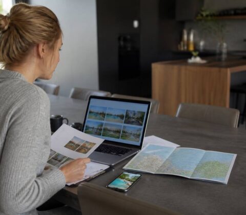 Persoon vergelijkt reisopties op laptop en smartphone met reisnotities op tafel, bij daglicht, fotorealistisch.
